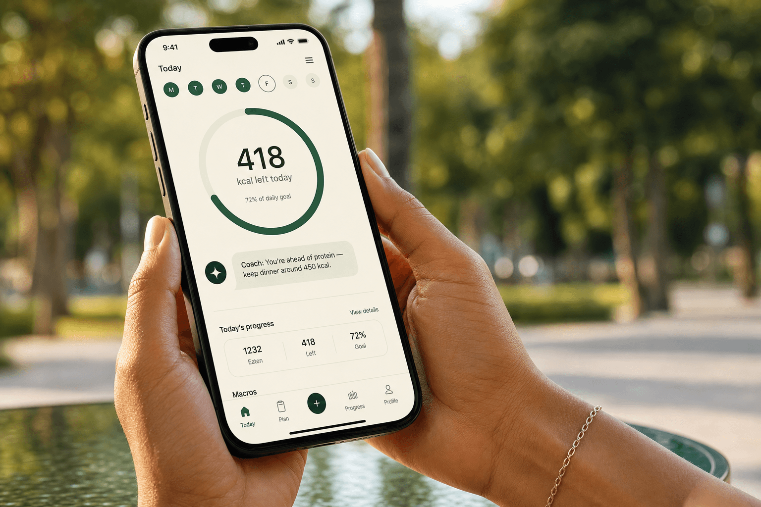 Two hands holding a phone running the GoodFitCoach AI app: the home screen shows the M-T-W-T-F-S-S day strip with Friday highlighted, the emerald calorie ring reads 418 kcal left today (72% of daily goal), a coach bubble suggests dinner around 450 kcal, and the Today's progress card breaks down 1232 eaten / 418 left / 72% goal — all over a softly blurred green park background.